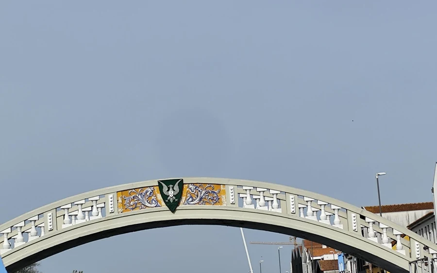 A white painted footbridge arch with a coat of arms on it, grey sky behind