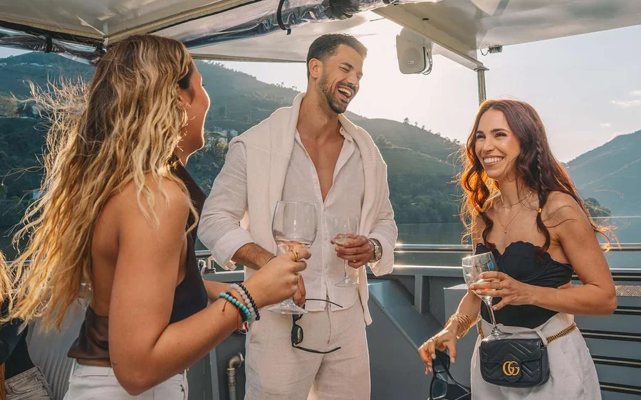 Three people with drinks on the open deck of a boat, green hills behind them
