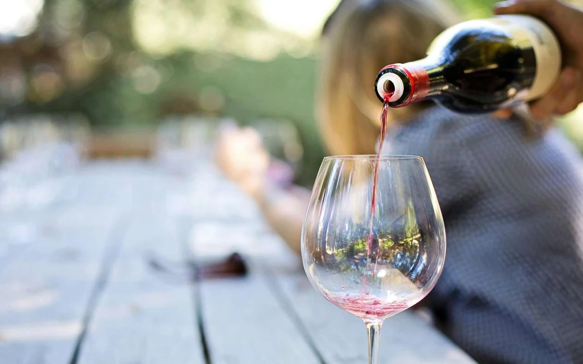 Red wine being poured from a dark bottle into a large glass on a weathered wooden table outdoors, with green leaves blurred behind